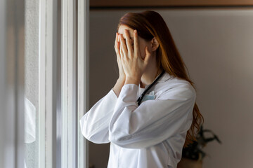 Emotionally stressed young female doctor with head in hands standing by window
