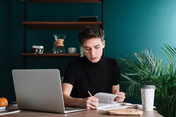 Male student writing in book while sitting with laptop at table doing homework