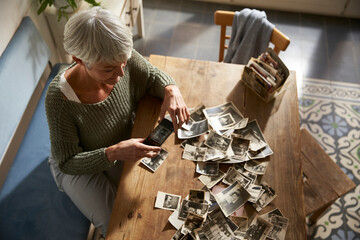 Senior woman sitting at table at home photographing old photos with smartphone
