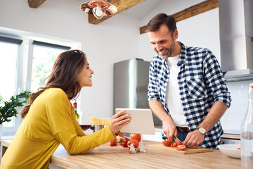 Affectionate couple in kitchen, preparing spaghetti toghether, using digital tablet - obrazy, fototapety, plakaty