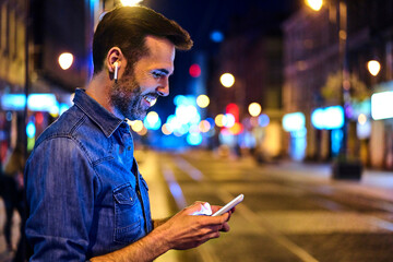 Man with wireless headphones using smartphone in the city at night - obrazy, fototapety, plakaty