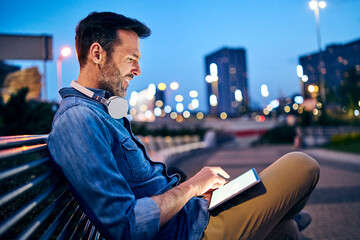 Man using tablet while sitting on bench out in the city durign evening