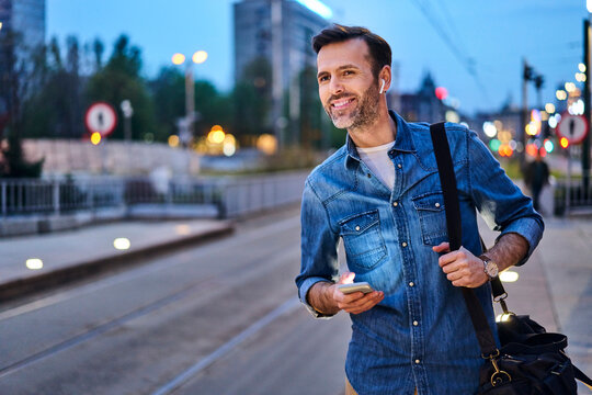 Smiling man with wireless headphones and smartphone waiting at tram stop during evening commute after work