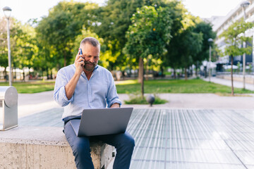 Businessman using laptop while talking on mobile phone at office park