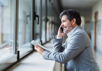 Smiling male professor talking on phone while looking through window in corridor at university