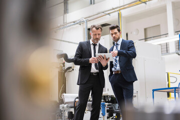 Two businessmen sharing tablet in factory shop floor - obrazy, fototapety, plakaty