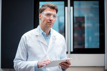 Portrait of man wearing lab coat and safety goggles holding tablet at machine