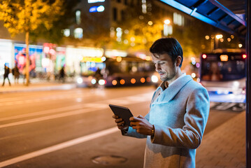 Businessman with digital tablet standing at a bus stop at night