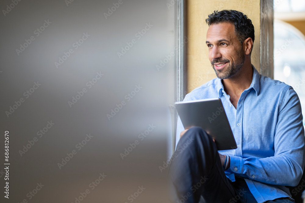 Wall mural Smiling businessman sitting at the window using tablet