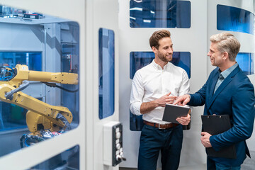 Two businessmen talking at robot in a modern factory