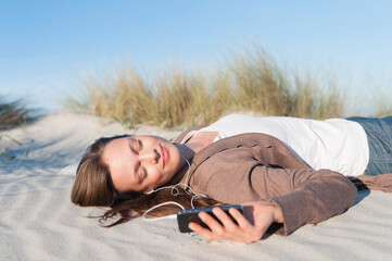 Portrait of woman lying on beach dune listening music with earphones and smartphone, Sardinia, Italy