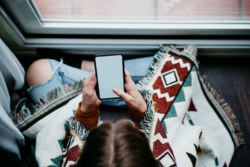 Girl with blanket using mobile phone while sitting by window at home
