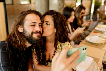 Man taking selfie while woman kissing him at restaurant