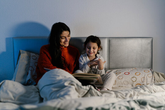 Happy mother and her little daughter sitting together on bed watching picture book