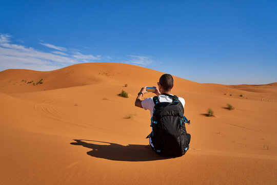 Morocco, man with backpack sitting on desert dune taking picture with smartphone