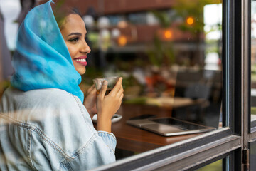 Young woman wearing turquoise hijab and drinking tea in a cafe, looking through window