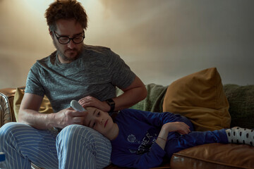 Man holding thermometer on sick daughter's forehead in living room at home - obrazy, fototapety, plakaty