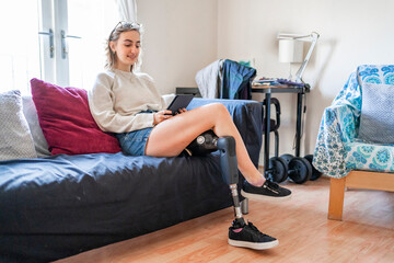 Young woman with leg prosthesis sitting on couch at home reading e-book