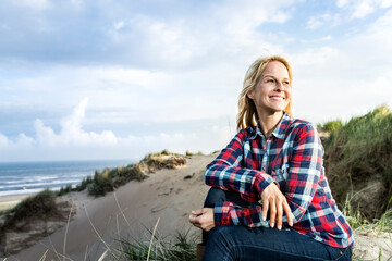 Smiling woman sitting in dunes - obrazy, fototapety, plakaty