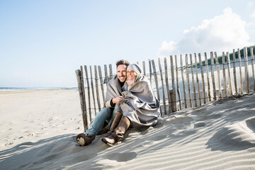 Smiling couple wrapped in blanket on the beach - obrazy, fototapety, plakaty