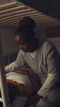 Medium vertical shot of young African American boy sleeping in bed at night, mom in hospital uniform coming in, tucking duvet, kissing him on cheek, then leaving and closing bedroom door