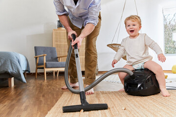 Low section of man cleaning carpet while cute baby boy sitting on vacuum cleaner in living room at home - Powered by Adobe