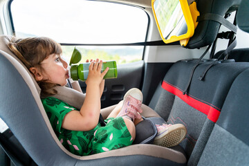 Toddler girl sitting on a car seat with a mirror drinking water