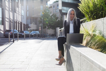 Young businesswoman sitting on a wall in the city using laptop - obrazy, fototapety, plakaty