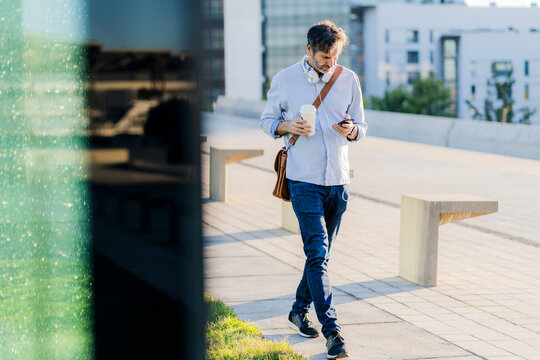Mature man with takeaway coffee and headphones using cell phone in the city