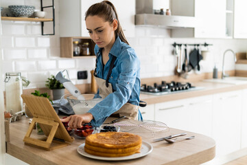 Young woman preparing a cream cake, using tablet - obrazy, fototapety, plakaty