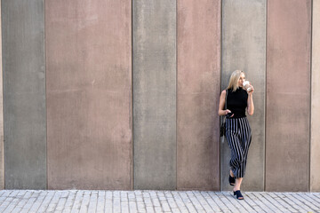 Young blond woman with cell phone leaning against wall drinking coffee to go