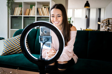 Smiling beautiful female influencer sitting on sofa while vlogging through smart phone at home