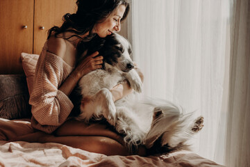 Happy woman sitting on bed at home stroking her dog - obrazy, fototapety, plakaty