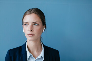 Portrait of a young businesswoman against blue background, wearing ear buds
