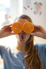 Playful young woman covering her eyes with oranges at home