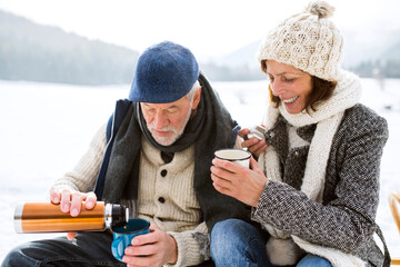 Senior couple having a break with hot beverages in snow-covered winter landscape - obrazy, fototapety, plakaty