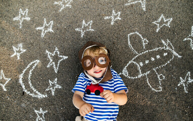 Portrait of smiling toddler wearing pilot hat and goggles lying on asphalt painted with airplane,...