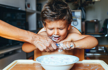 Little boy eating yogurt at home, hand of woman on the spoon