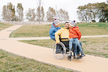 Playful man with excited sons riding wheelchair on footpath in park