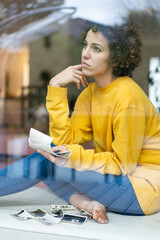 Serious woman sitting behind window at home holding photos