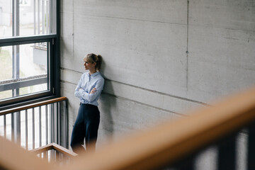 Successful businesswoman leaning on wall, looking out of window, with arms crossed