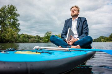 Businessman with laptop sitting on SUP board on a lake