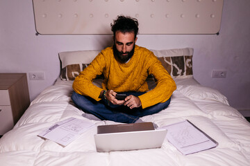 Young man working in bed at home with cell phone, laptop and documents