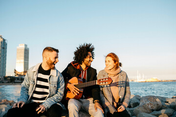 Three happy friends with guitar sitting outdoors at the coast sunset