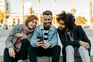 Three happy friends sitting outdoors looking at cell phone