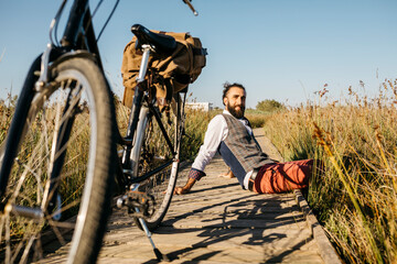 Well dressed man sitting on a wooden walkway in the countryside next to a bike