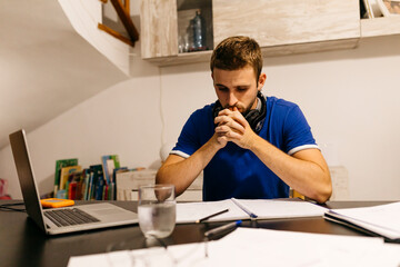 Dedicated young male student sitting with hands clasped while doing homework at table