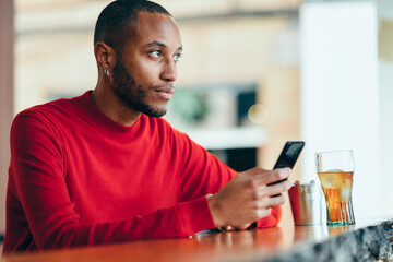 Pensive young man wearing red pullover at counter of a bar with soft drink and mobile phone