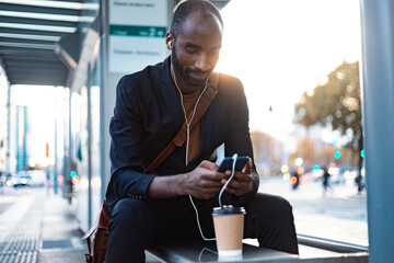 Young businessman sitting at tram stop in the evening using earphones and smartphone - obrazy, fototapety, plakaty