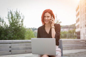 Young woman sitting on bench with her laptop, making a phone call